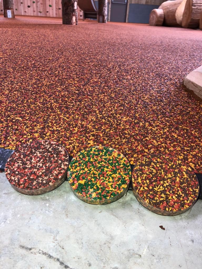 A photograph of three circular samples showing colours aggregates of RubbaSmart wet pour rubber in front of a pre-laid red, yellow and brown rubber surface. One sample is red, brown, and peach, the next is green, yellow and red, and the last is red, yellow and brown