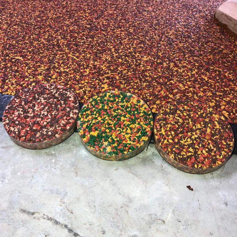 Three circular samples showing colours aggregates of RubbaSmart wet pour rubber in front of a pre-laid red, yellow and brown rubber surface. One sample is red, brown, and peach, the next is green, yellow and red, and the last is red, yellow and brown