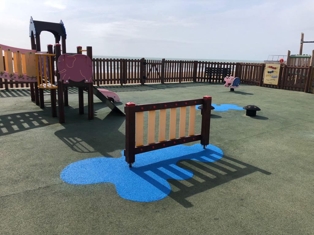 An image of RubbaSmart wet pour rubber playground surfacing being used on a playground. Over green artificial surfacing, blue wet pour is used to create a puddle shaped design beneath some playground equipment.