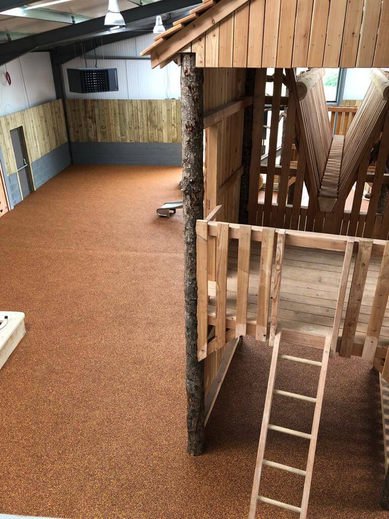 Wet Pour Rubber installation. The brown rubber covers a playground surface, around wooden play structures and a workers is laying and smoothing out a final section