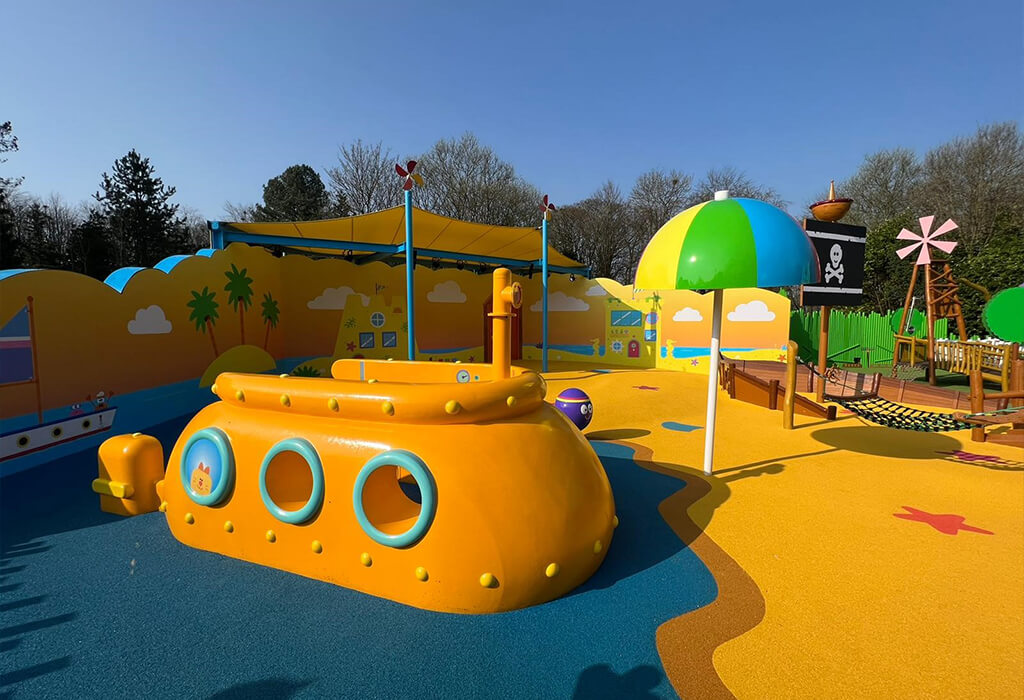 Alt-Tag- Yellow, orange, and dark blue coloured RubbaSmart wet pour rubber surfacing used to create a beach and sea design on a children’s playground. Playground structures on this site include a yellow submarine, beach umbrella and pirate ship.