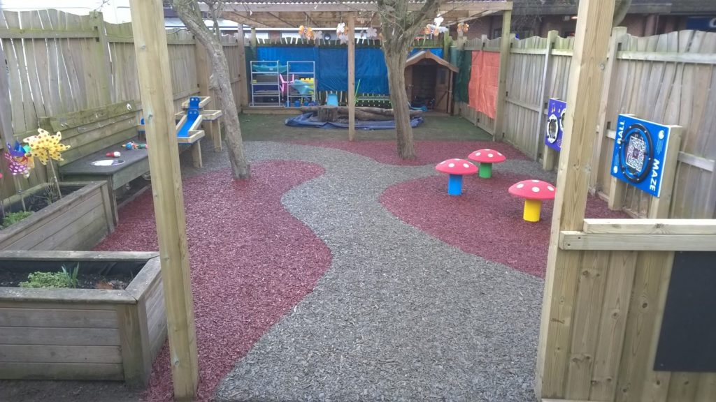 A school playground using our Jungle Mulch Rubber Mulch surfacing solution in the colours gray and pink in a unique and interest design fitting with mushroom shaped seats and wooden planters