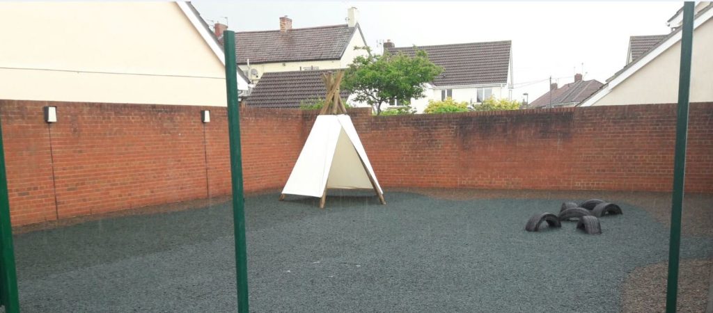 A playground with a wooden and cloth teepee and red brick wall, using our Jungle Mulch Rubber Mulch surfacing cover in blue and gray colours.