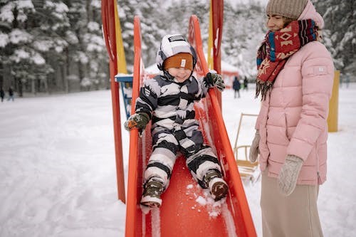 Child sliding down a red plastic slide in a snowy playground with their parent watching, both wrapped up warm in winter clothes