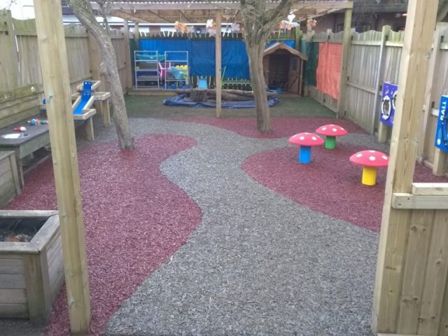 A school playground using our Jungle Mulch Rubber Mulch surfacing solution in the colours gray and pink in a unique and interest design fitting with mushroom shaped seats and wooden planters