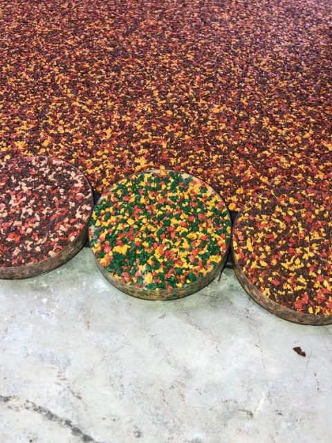 Three circular samples showing colours aggregates of RubbaSmart wet pour rubber in front of a pre-laid red, yellow and brown rubber surface. One sample is red, brown, and peach, the next is green, yellow and red, and the last is red, yellow and brown