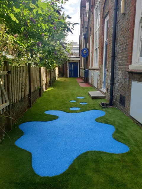 Artificial grass safety surface installation along a building with puddle shaped EPDM wear pad