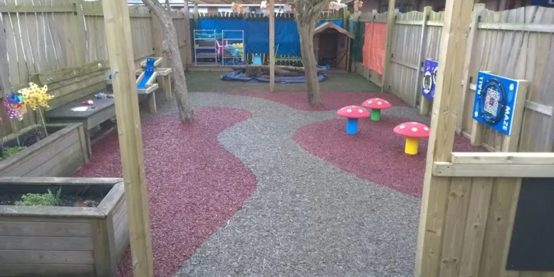 A school playground using our Jungle Mulch Rubber Mulch surfacing solution in the colours gray and pink in a unique and interest design fitting with mushroom shaped seats and wooden planters