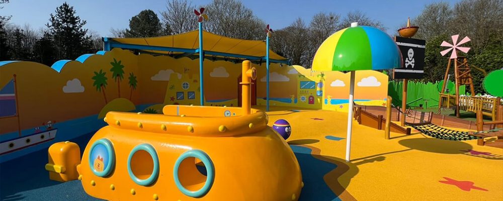 Alt-Tag- Yellow, orange, and dark blue coloured RubbaSmart wet pour rubber surfacing used to create a beach and sea design on a children’s playground. Playground structures on this site include a yellow submarine, beach umbrella and pirate ship.