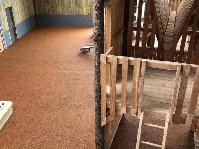 Wet Pour Rubber installation. The brown rubber covers a playground surface, around wooden play structures and a workers is laying and smoothing out a final section