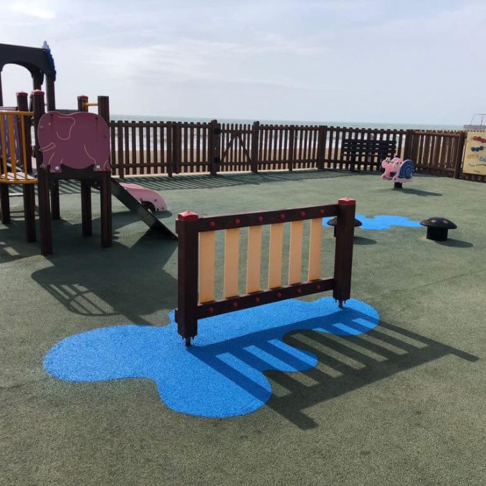 RubbaSmart wet pour rubber playground surfacing being used on a playground. Over green artificial surfacing, blue wet pour is used to create a puddle shaped design beneath some playground equipment